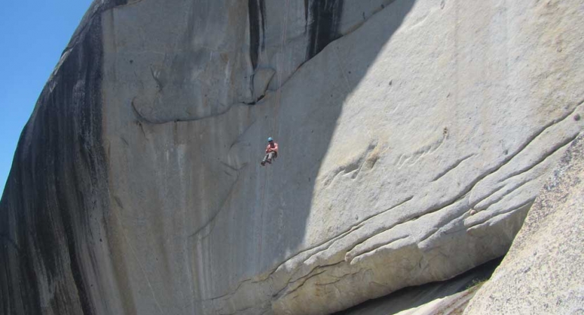 a person wearing safety gear is suspended by ropes mid air against the backdrop of a large smooth rock wall